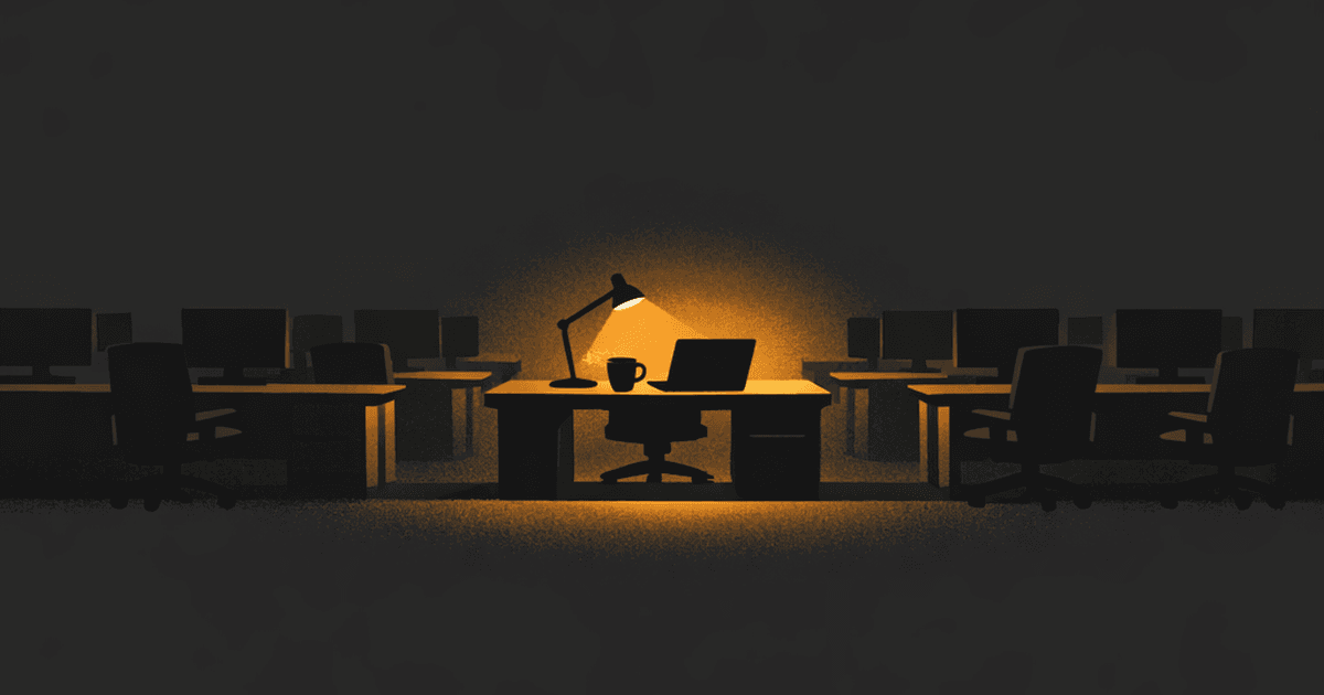 A single lit desk lamp in a dark open-plan office full of empty chairs and blank monitors — the quiet aftermath of a layoff wave